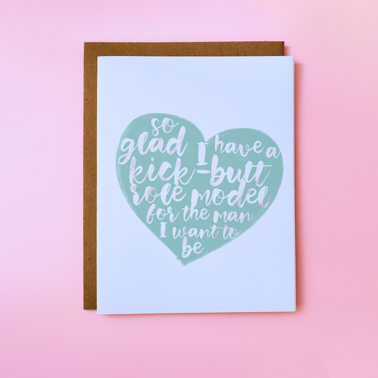 A LGBTQ Parent Card with a kraft envelope set on a pink background. This inclusive parent Day Card features a pgreen heart with text that reads 'So glad I have a kick-butt role model for the man I want to be'.