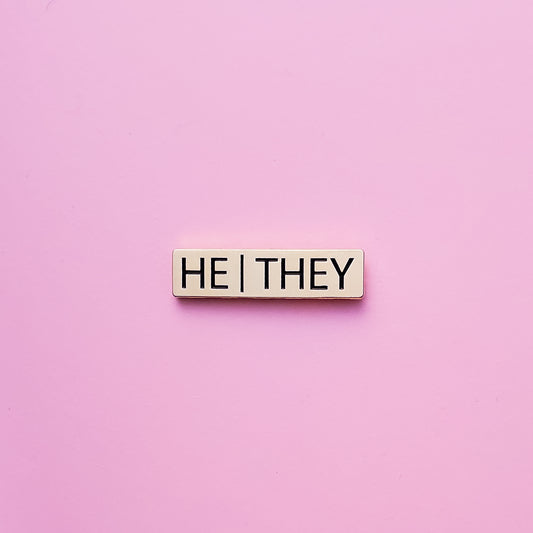 A Gold bar-shaped enamel pin with he they pronouns set on a pink background. This He They Pronoun Pin has black enamel set in a polished gold metal.He/They Pronoun Pin - LGBTQ Pride Pins | Little Rainbow Paper Co