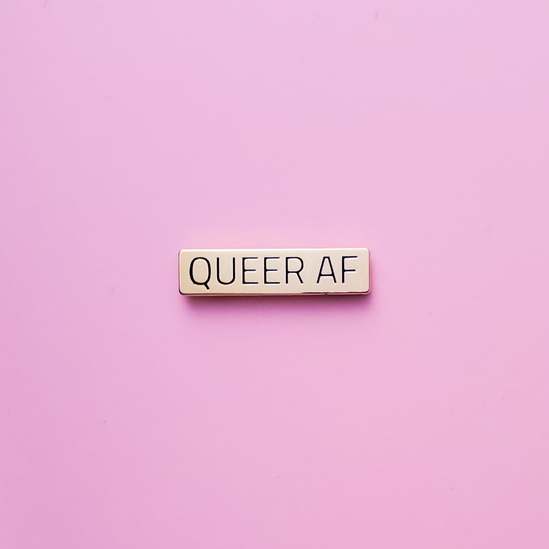 Gold bar-shaped enamel pin reads 'Queer AF' against a pink background. This LGBTQ pin is set in a polished gold color metal. Queer AF Pin - LGBTQ Pins For Sale | Little Rainbow Paper Co