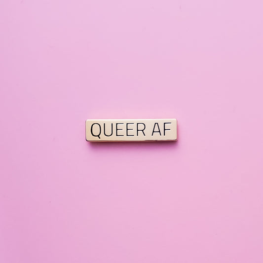 Gold bar-shaped enamel pin reads 'Queer AF' against a pink background. This LGBTQ pin is set in a polished gold color metal. Queer AF Pin - LGBTQ Pins For Sale | Little Rainbow Paper Co