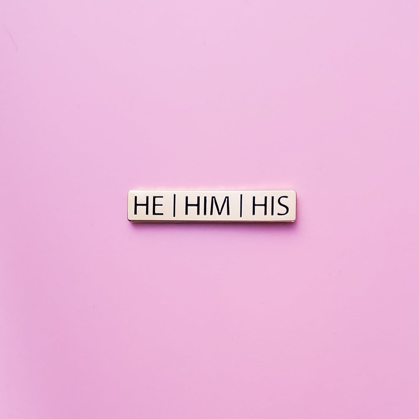 A gold bar shaped He Him His Pronoun Pin on a pink background. This He Him His Pronoun Pin has black enamel set in a polished gold metal. He/Him/His Pronoun Pin - LGBTQ Pride Pins Canada | Little Rainbow Paper Co