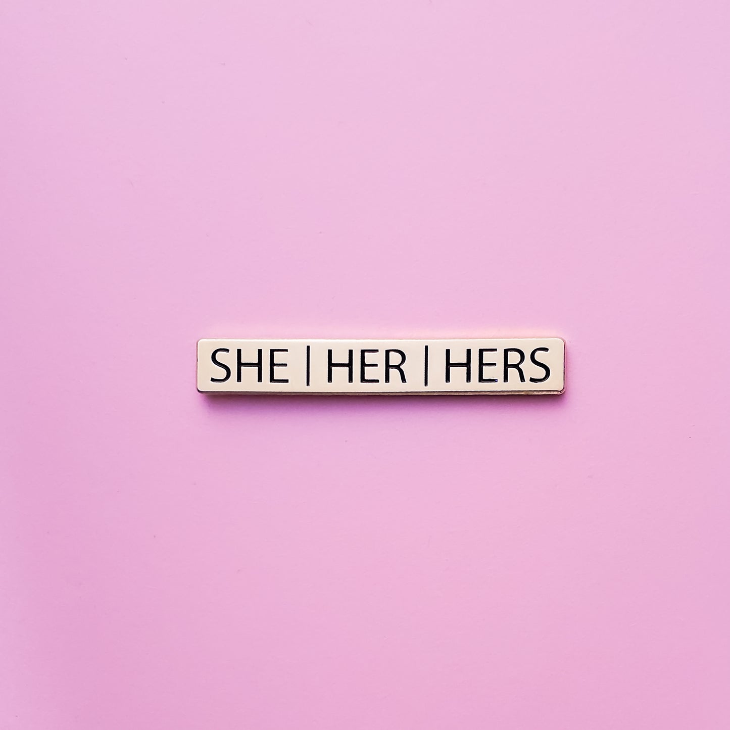A Gold bar-shaped enamel pin with she/her/hers pronouns set on a pink background. This She Her Hers Pronoun Pin has black enamel set in a polished gold metal. She/Her/Hers Pronoun Pin - LGBTQ Pride Pins | Little Rainbow Paper Co