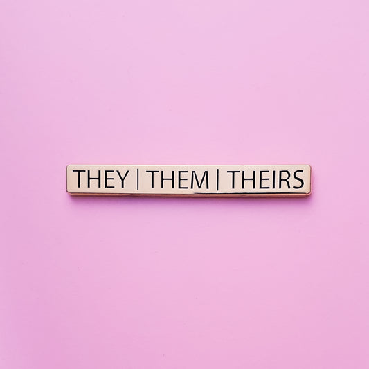 A Gold bar-shaped enamel pin with&nbsp;they them theirs pronouns set on a pink background. This They Them Theirs Pronoun Pin has black enamel set in a polished gold metal.. They/Them/Theirs Pronoun Pin For Sale - LGBTQ Accessories  | Little Rainbow Paper Co