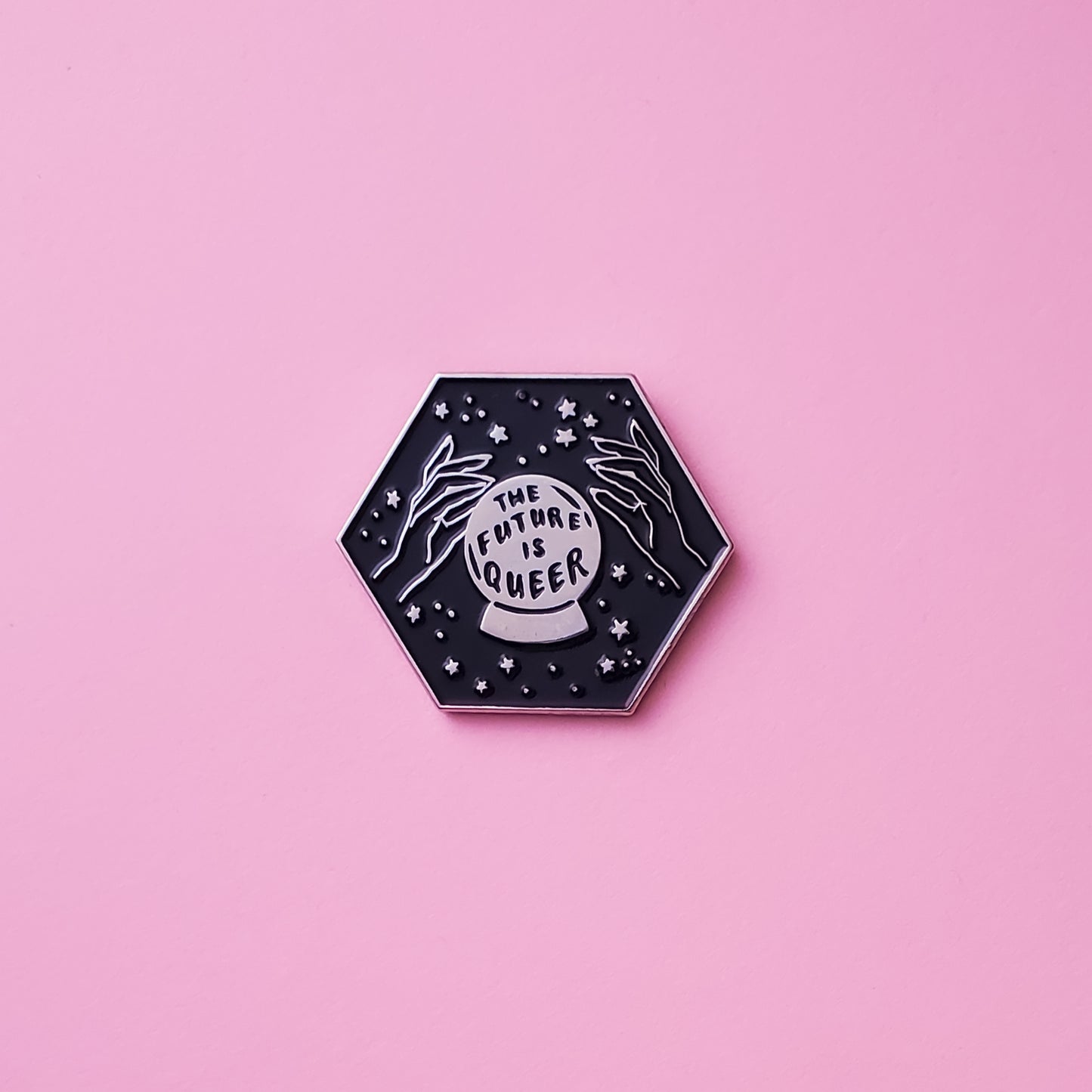 Hexagonal shaped pride pin on a pink background. This LGBTQ pin features two hands, stars, and a crystal ball that reads ' The Future is Queer' against black enamel. Future is Queer Pin For Sale - LGBTQ Accessories  | Little Rainbow Paper Co