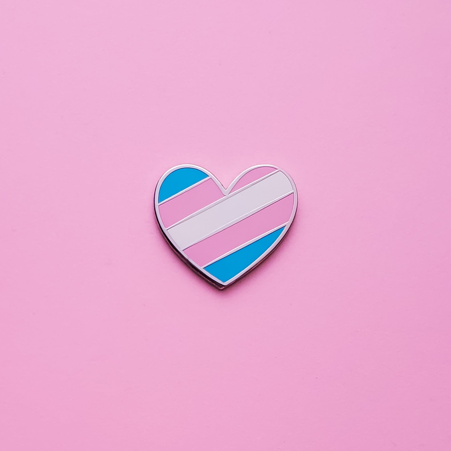 Heart-shaped Trans Pride Pin on a pink background. This Trans pin is has vibrant Transgender pride colours set in a polished nickle metal. Trans pride pin colours are blue, pink and white. Trans Heart Pin - LGBTQ Pins Canada | Little Rainbow Paper Co