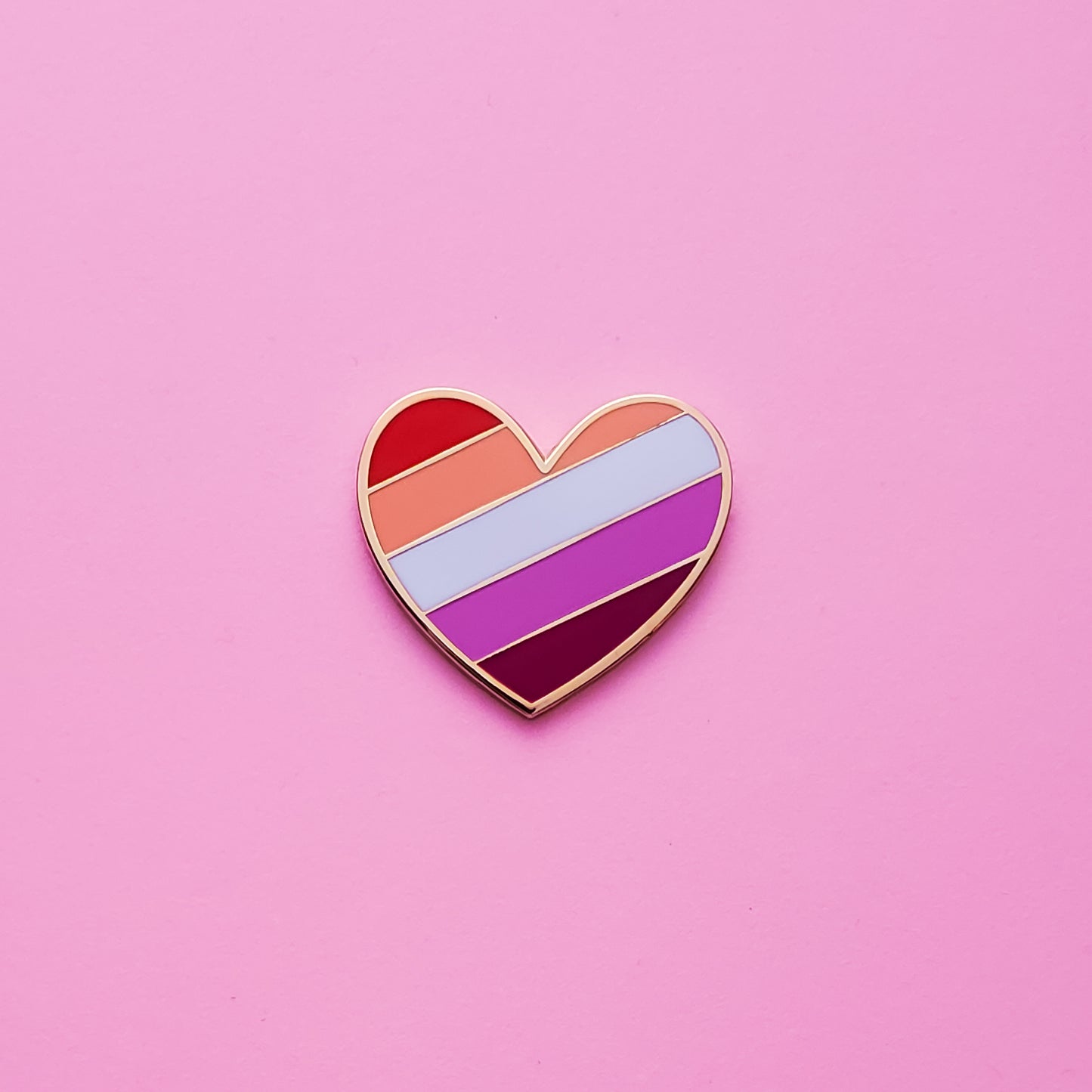 Heart-shaped Lesbian Pride Pin on a pink background. This lesbian pin is has vibrant Lesbian pride colours set in a polished gold metal. Lesbian pride pin colours are red, orange, white, light purple, dark purple. Lesbian Heart Pin For Sale - LGBTQ Accessories  | Little Rainbow Paper Co
