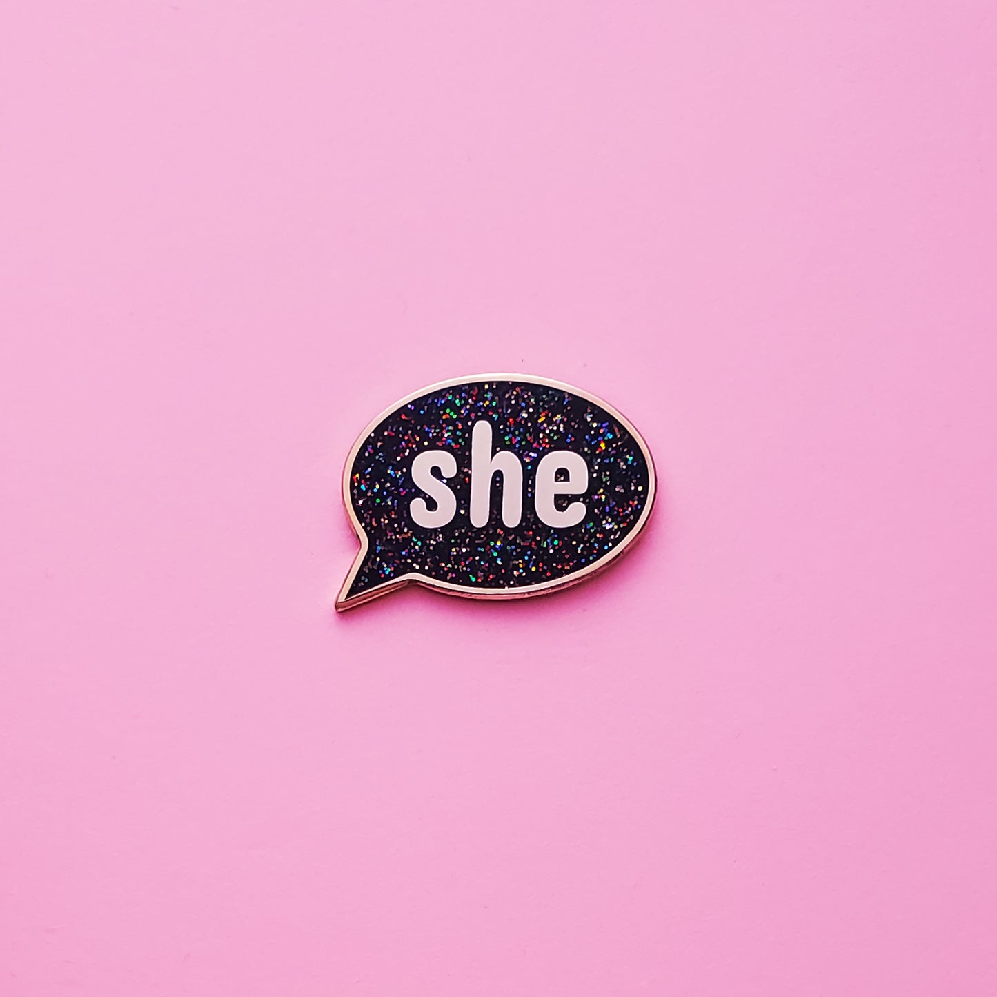 A Speech-bubble shaped She Pronoun Pin on a pink background. This She Pronoun Pin has black  glitter enamel set in a polished gold metal. She Pronoun Pin - LGBTQ Pins Canada | Little Rainbow Paper Co