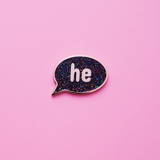 A Speech-bubble shaped He Pronoun Pin on a pink background. This He Pronoun Pin has black glitter enamel set in a polished gold metal. - LGBTQ Pride Pins Canada | Little Rainbow Paper Co