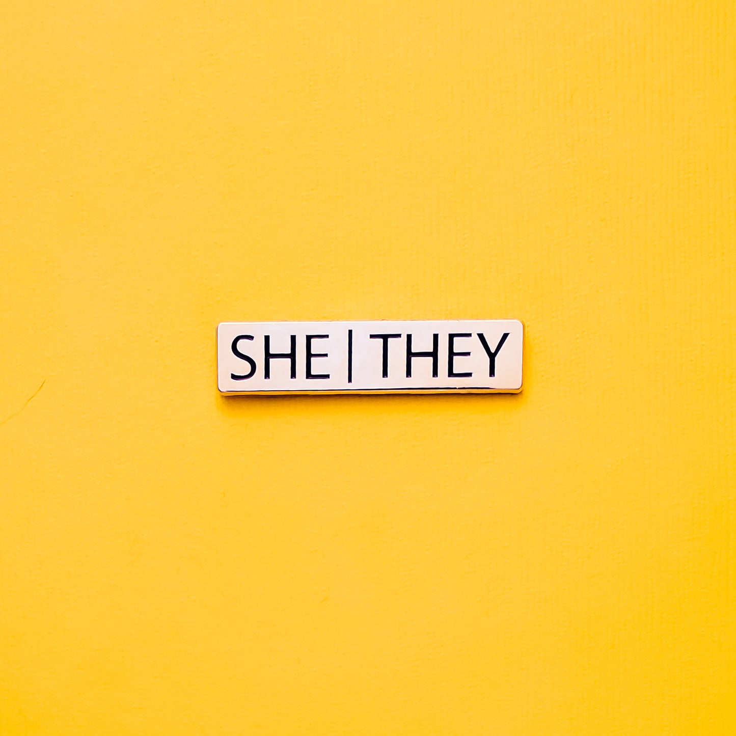 A Gold bar-shaped enamel pin with she they pronouns set on a yellow background. This He They Pronoun Pin has black enamel set in a polished gold metal.She/They Pronoun Pin - LGBTQ Pride Pins | Little Rainbow Paper Co