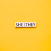 A Gold bar-shaped enamel pin with she they pronouns set on a yellow background. This He They Pronoun Pin has black enamel set in a polished gold metal.She/They Pronoun Pin - LGBTQ Pride Pins | Little Rainbow Paper Co