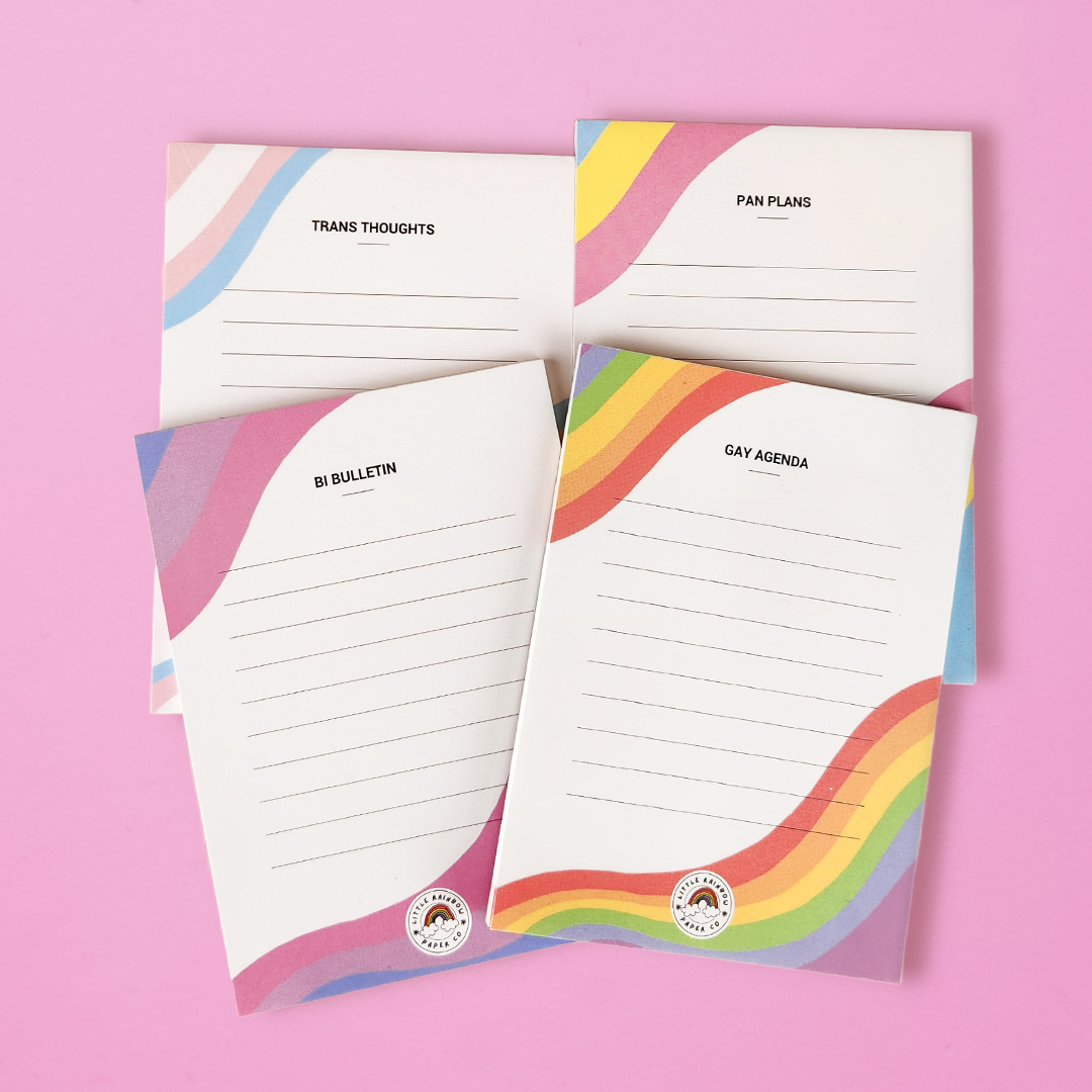 Four LGTBQ notepads on a pink background. A trans pride notepad reads trans thoughts. A Pan pride notepad reads pan plans. A Bi pride notepad reads Bi Bulletin. A gay pride notepad reads gay agenda.