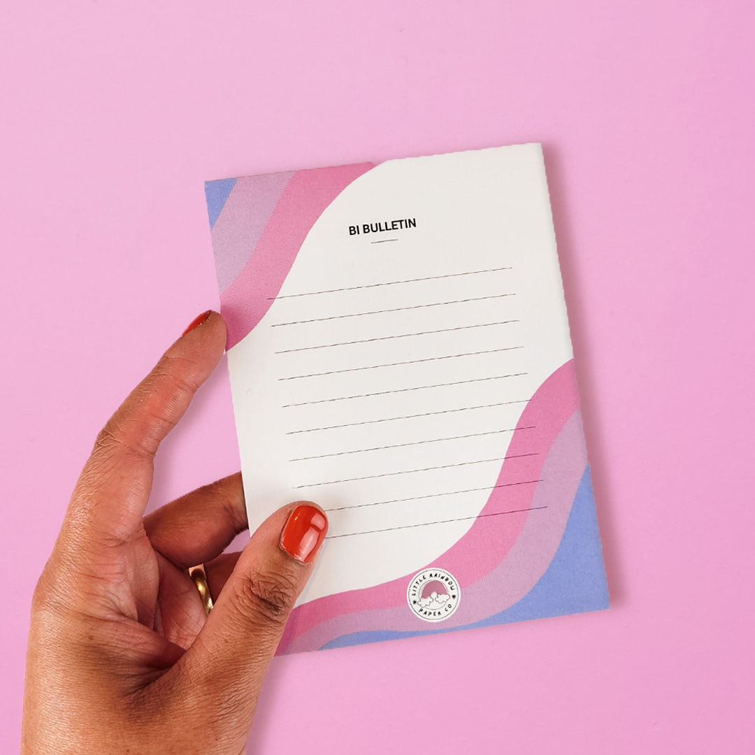 A hand holds a LGTBQ notepad with the bi pride flag colors printed on the front. Text reads bi bulletin and has lines for writing. A cute bisexual notepad