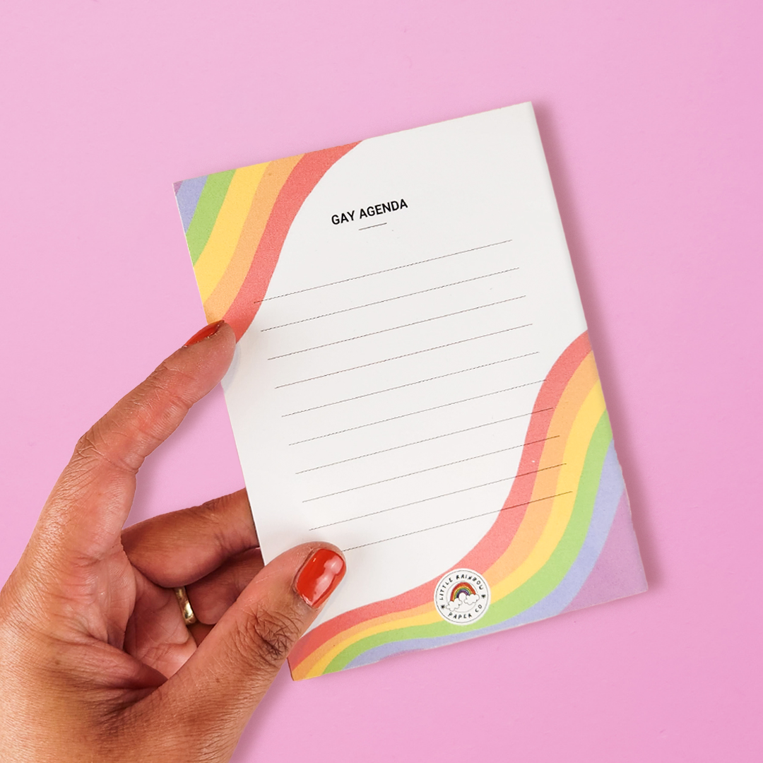 A hand holds a LGTBQ notepad with the gay pride flag colors printed on the front. Text reads gay agenda and has lines for writing. A cute gay pride notepad