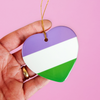 Hand holding a ceramic heart shaped LGBTQ ornament with the gender queer pride colors on a pink background. Heart-shaped gender queer pride ornament