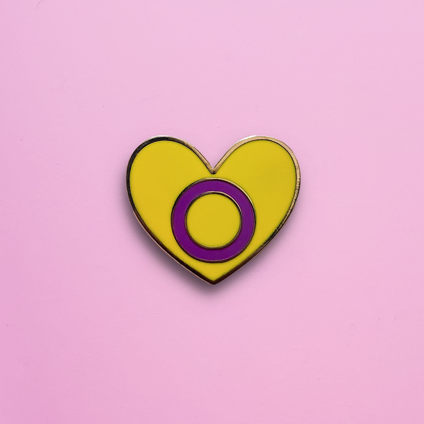 A Heart-shaped Intersex Pride Pin on a pink background. This Intersex pride pin is has vibrant Intersex pride colours set in a polished gold metal. Intersex pride pin colours are yellow with a  purple circle. Intersex Heart Pin - LGBTQ Pride Pins Canada | Little Rainbow Paper Co