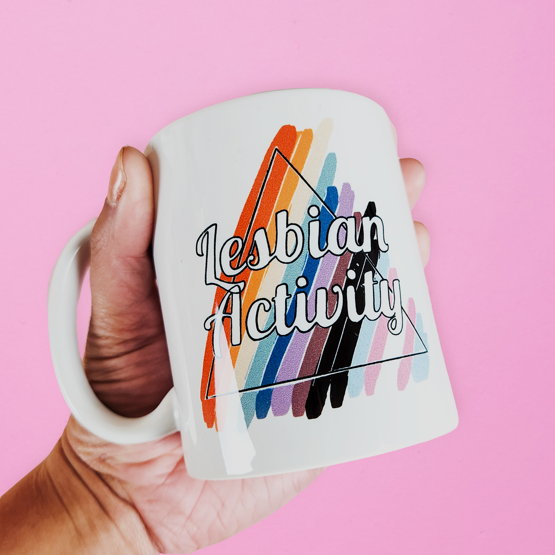 Hand holding LGBTQ mug that reads Lesbian Activity over progress pride colors. Cute lesbian mug on pink background