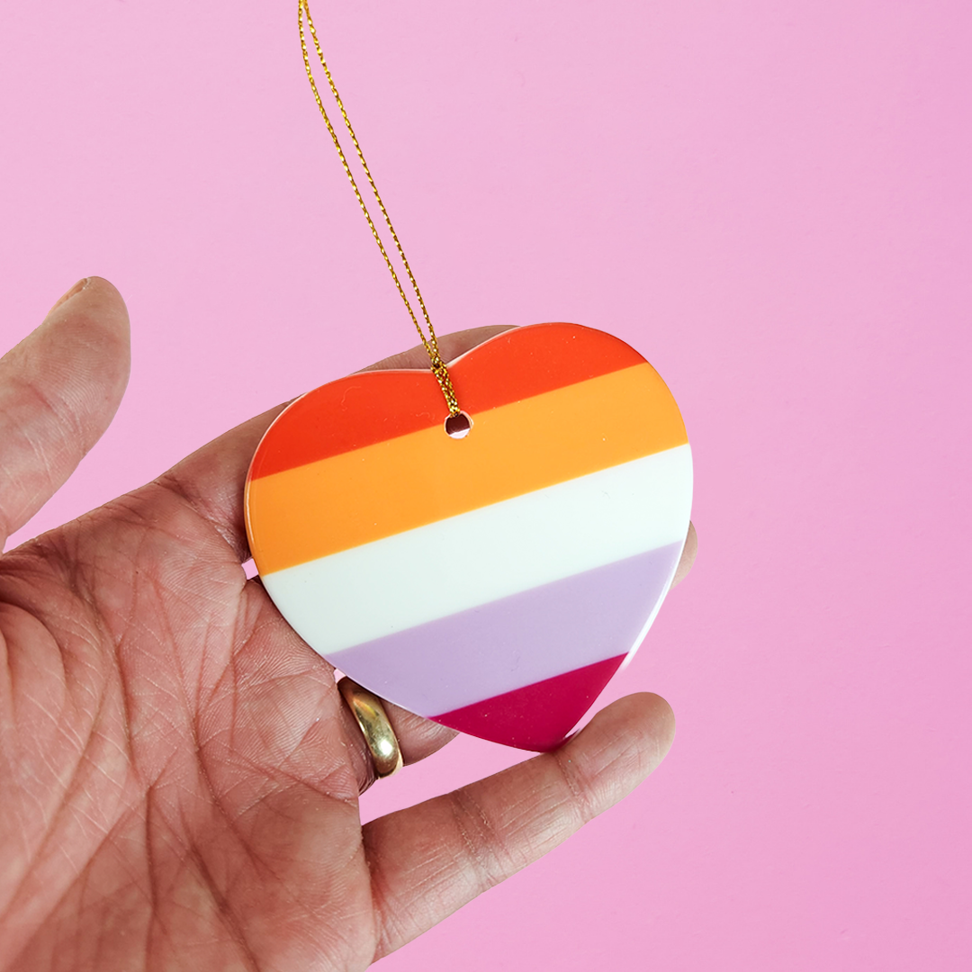 Hand holding a ceramic heart shaped LGBTQ ornament with the lesbian pride colors on a pink background. Heart-shaped lesbian pride ornament