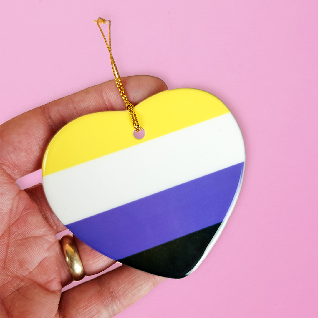 Hand holding a ceramic heart shaped LGBTQ ornament with the non-binary pride colors on a pink background. Heart-shaped non-binary pride ornament
