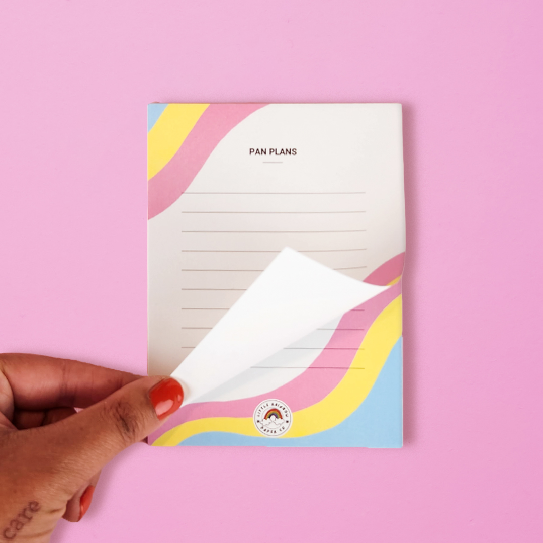 A hand flips a page on a LGTBQ notepad with the pan pride flag colors printed on the front. Text reads pan plans and has lines for writing. A cute pansexual pride notepad