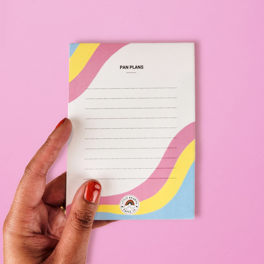 A hand holds a LGTBQ notepad with the pan pride flag colors printed on the front. Text reads pan plans and has lines for writing. A cute pansexual pride notepad