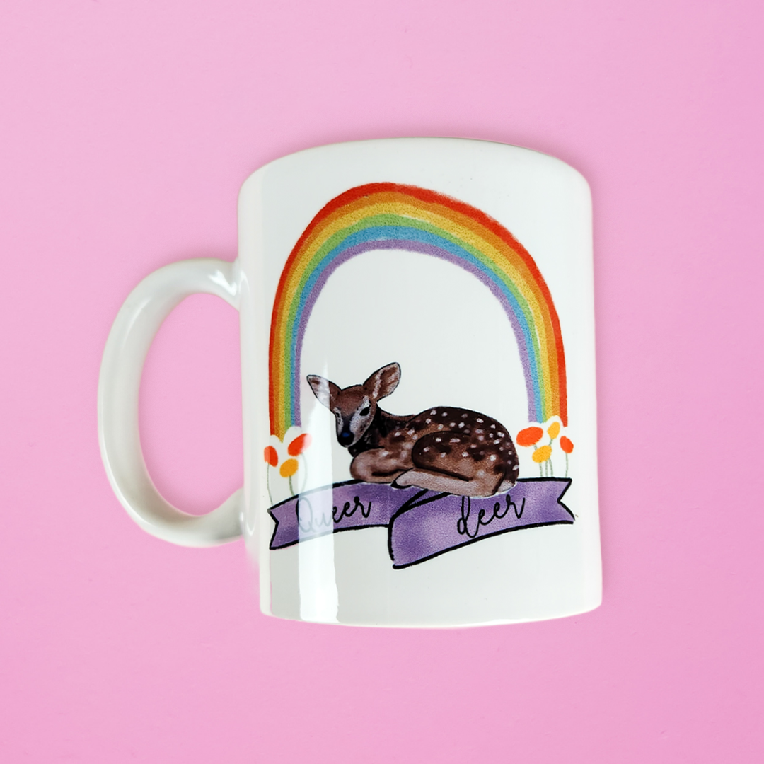 LGBTQ mug that has a deer sitting underneath a rainbow with a banner reading queer deer. Cute gay mug on a pink background