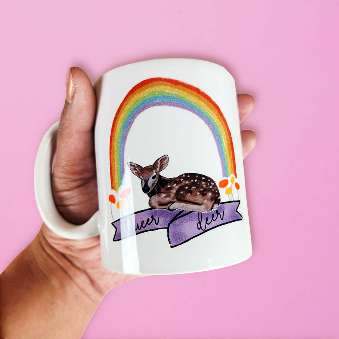 hand holding LGBTQ mug that has a deer sitting underneath a rainbow with a banner reading queer deer. Cute gay mug on a pink background