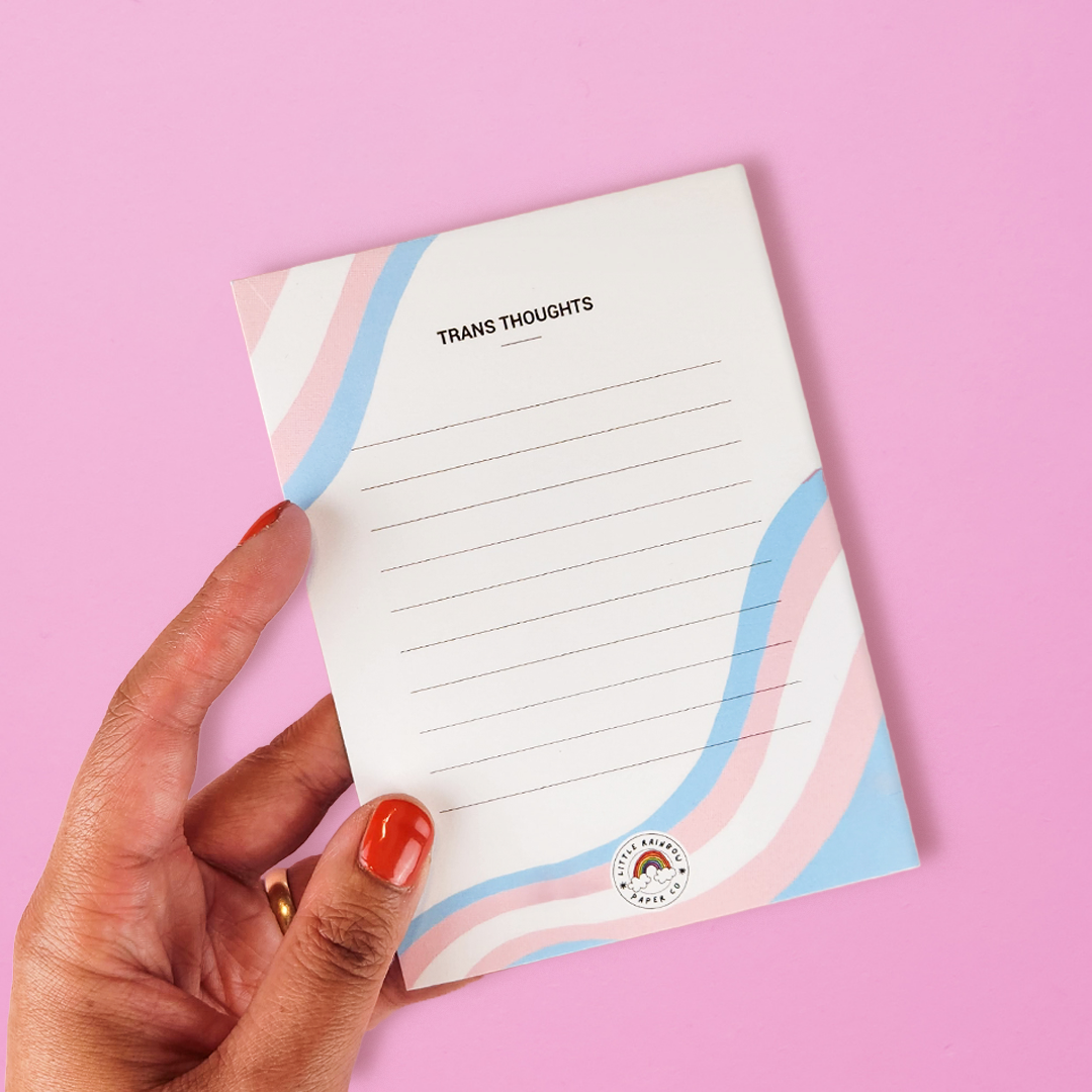 A hand holds a LGTBQ notepad with the trans pride flag colors printed on the front. Text reads trans thoughts and has lines for writing. A cute trans pride notepad