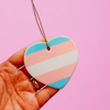 Hand holding a ceramic heart shaped LGBTQ ornament with the trans pride colors on a pink background. Heart-shaped trans pride ornament