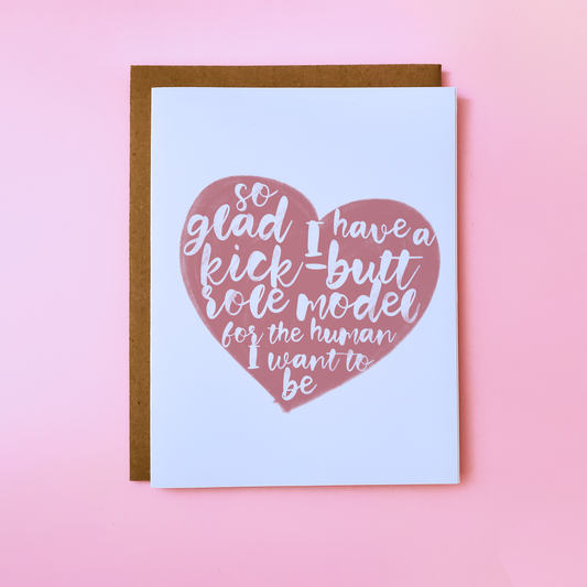 A LGBTQ Parent Card with a kraft envelope set on a pink background. This inclusive parent Day Card features a pink heart with text that reads 'So glad I have a kick-butt role model for the human I want to be'.