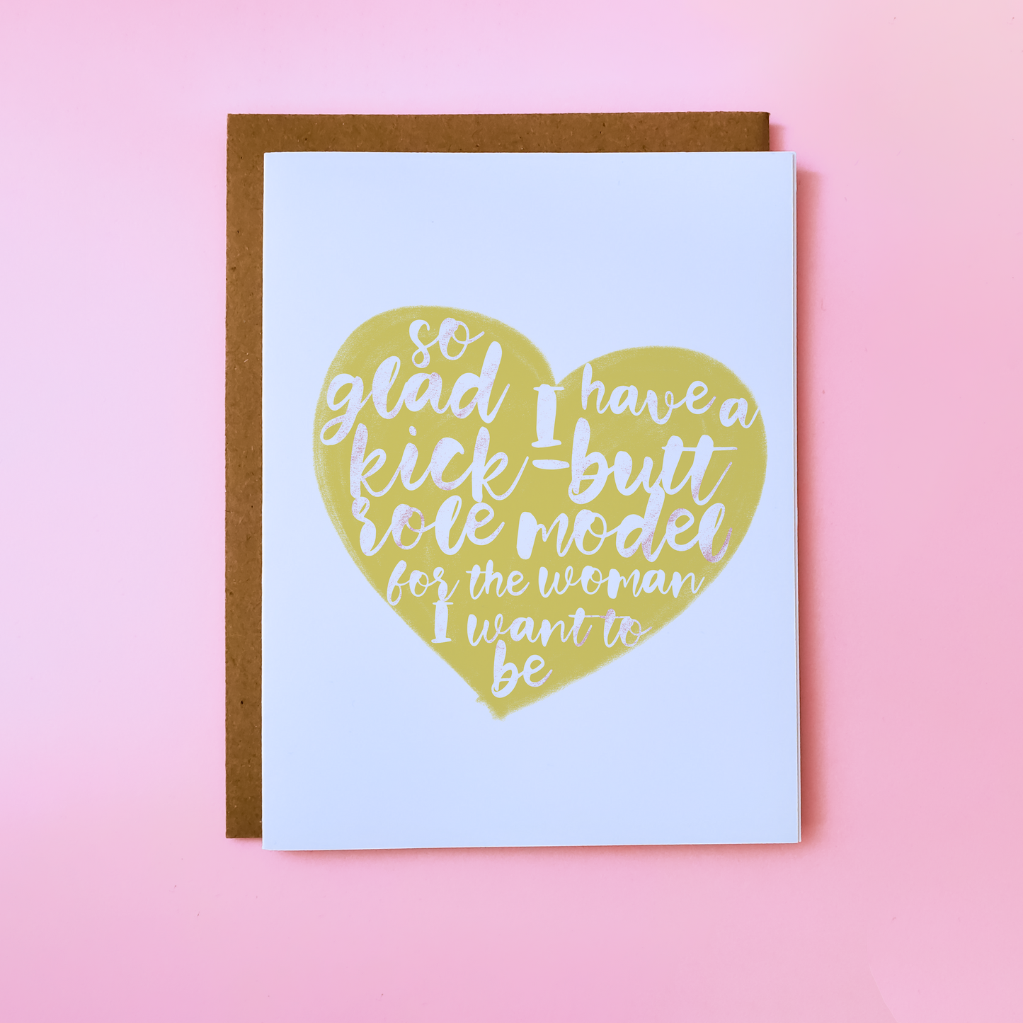 A LGBTQ Parent Card with a kraft envelope set on a pink background. This inclusive parent Day Card features a yellow heart with text that reads 'So glad I have a kick-butt role model for the woman I want to be'.