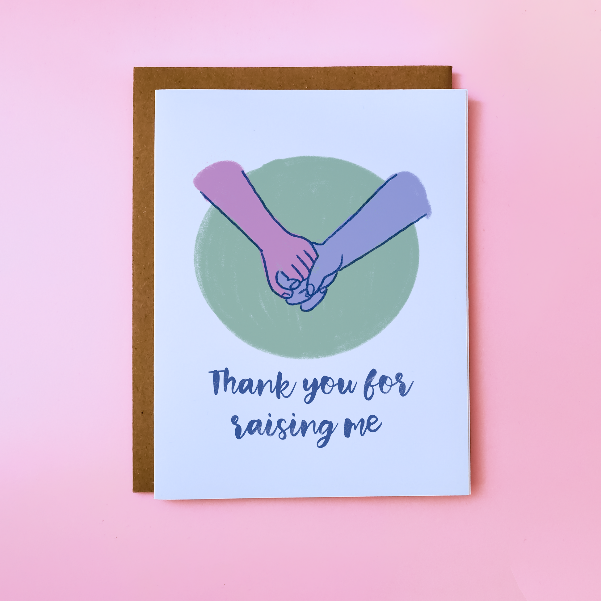 A LGBTQ Parent Card with a kraft envelope set on a pink background. This inclusive Parent Card features hands holding on a green background and reads 'Thank You For Raising Me'.
